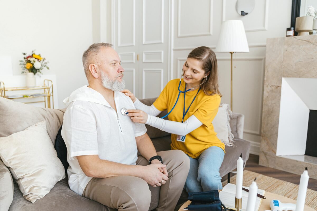 A nurse uses a stethoscope for a home check-up on a senior adult in a cozy living room.